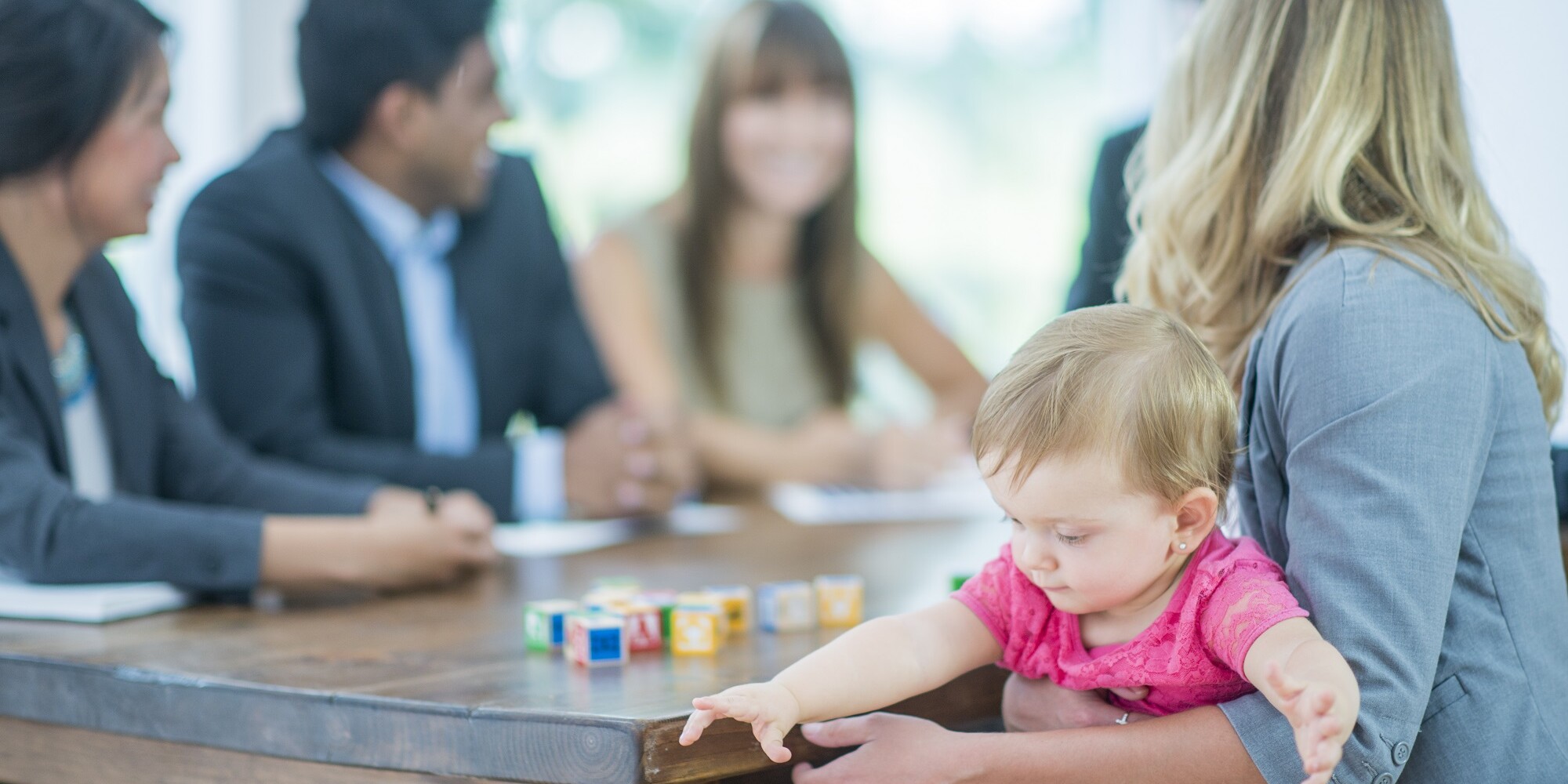 A multi-ethnic group of adults business-people are indoors in a board room. They are wearing formal clothing. A Caucasian woman is holding her baby during a meeting. The baby is playing with blocks.