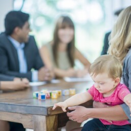 A multi-ethnic group of adults business-people are indoors in a board room. They are wearing formal clothing. A Caucasian woman is holding her baby during a meeting. The baby is playing with blocks.